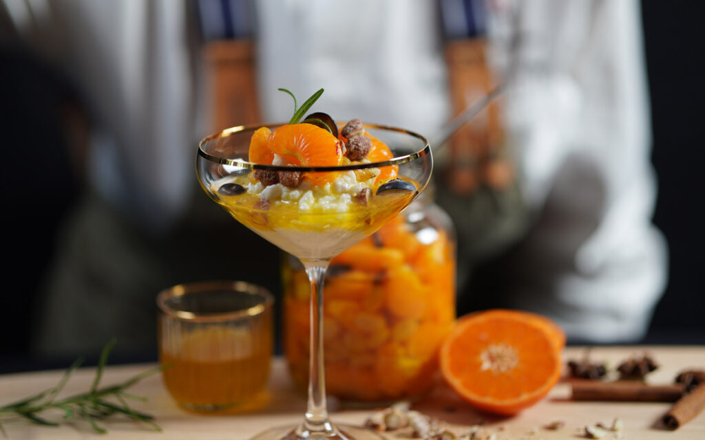 Dessert med risgröt, apelsin, clementin, kanel och stjärnanis. Serverad i högt glas. I bakgrunden syns en suddig kock med förkläde.